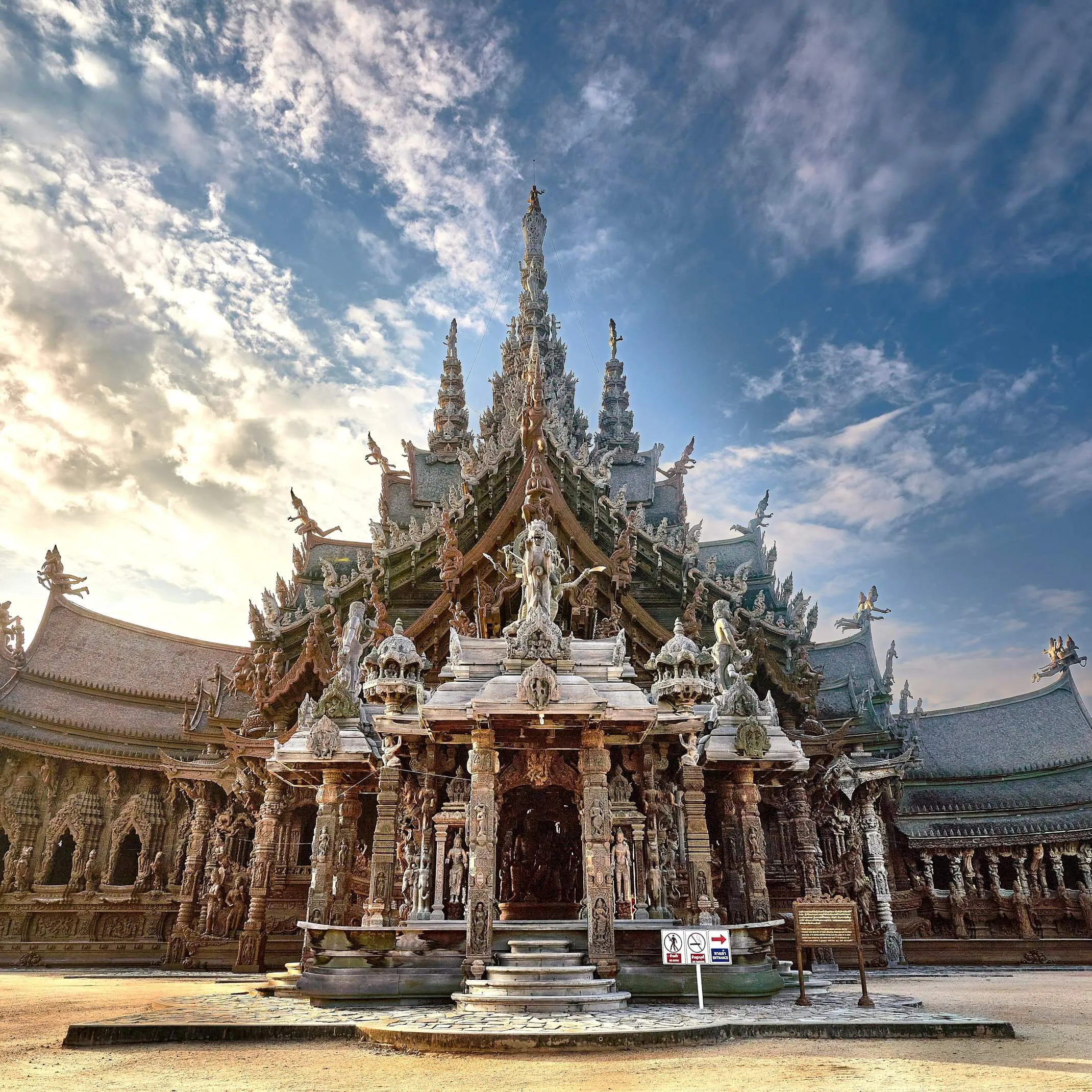 Front entrance of the Sanctuary of Truth: steps up to a carved wooden porch under tiered roofs and a tall central spire, against a blue sky with clouds